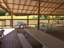 Shaded white picnic tables in an open-air pavilion with metal roof and wood-plank flooring. Sun-drenched  green grass in the background.